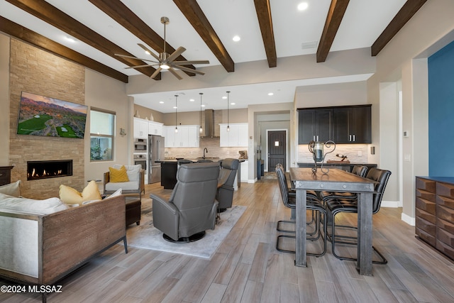 living room with light wood-type flooring, ceiling fan, sink, beam ceiling, and a stone fireplace