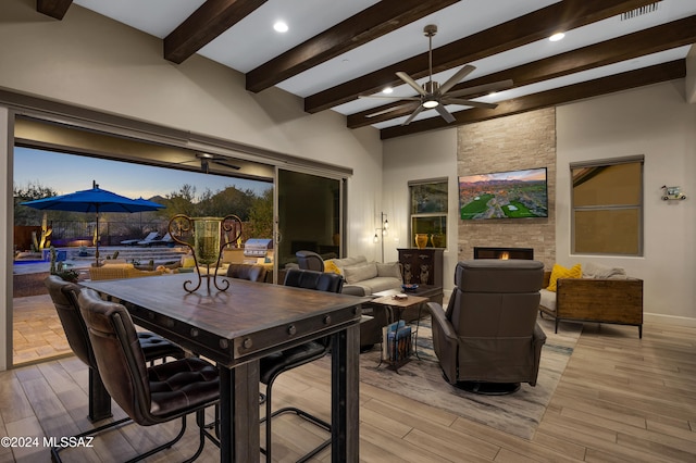 dining space featuring beamed ceiling, ceiling fan, a stone fireplace, and light wood-type flooring