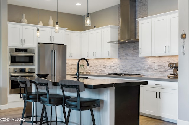 kitchen featuring appliances with stainless steel finishes, sink, wall chimney range hood, a center island with sink, and white cabinets