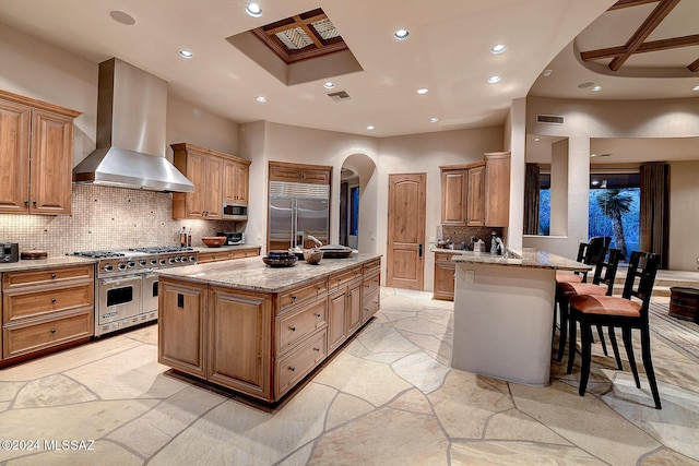 kitchen featuring built in appliances, a kitchen bar, a center island with sink, and wall chimney range hood