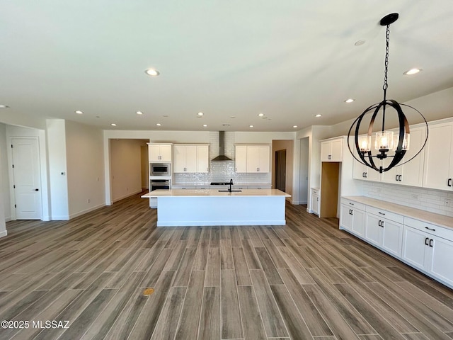 kitchen with white cabinets, light countertops, wall chimney range hood, appliances with stainless steel finishes, and an island with sink