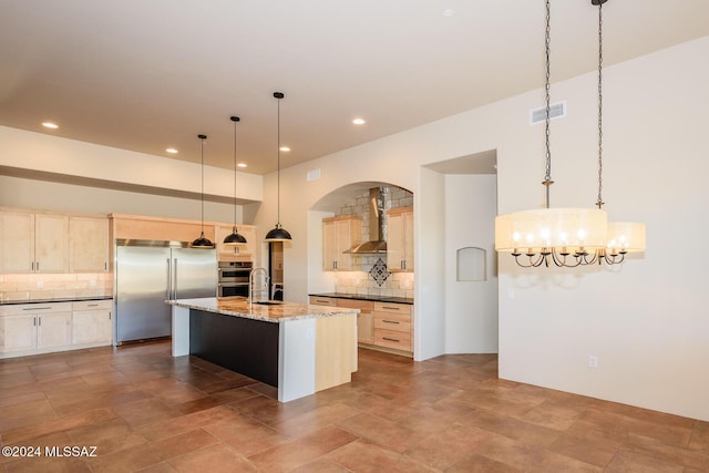 kitchen featuring appliances with stainless steel finishes, decorative light fixtures, dark stone countertops, a kitchen island with sink, and wall chimney exhaust hood