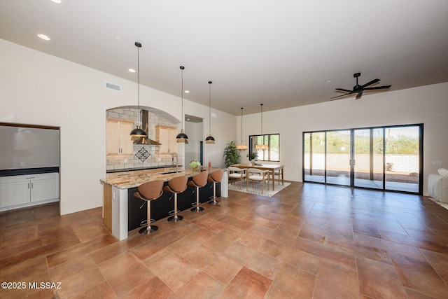 kitchen featuring a breakfast bar, pendant lighting, light stone countertops, a kitchen island with sink, and wall chimney range hood