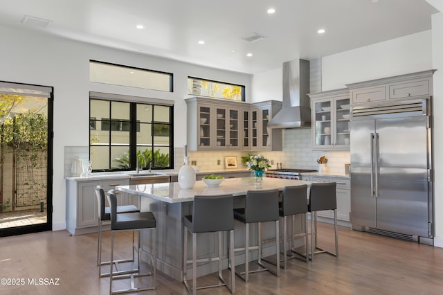 kitchen featuring built in refrigerator, wall chimney range hood, sink, light stone counters, and gray cabinetry