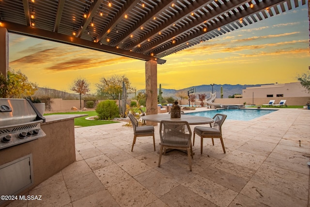 patio terrace at dusk featuring a mountain view, a fenced in pool, grilling area, an outdoor kitchen, and a pergola