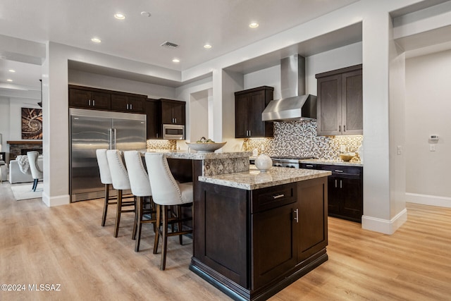 kitchen featuring a center island, built in appliances, wall chimney exhaust hood, light stone countertops, and light hardwood / wood-style floors