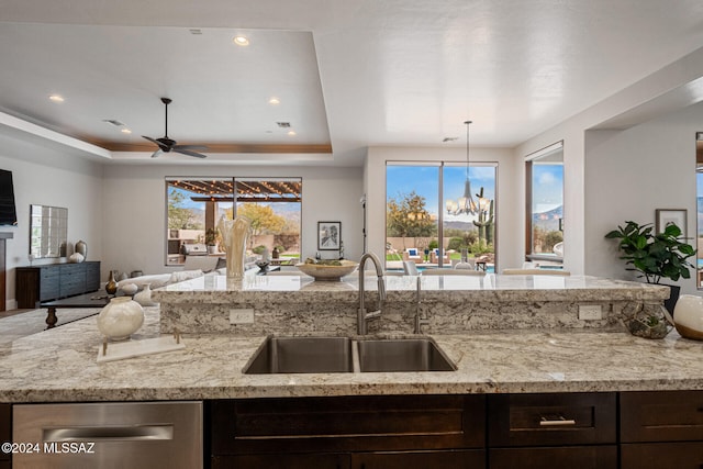 kitchen with ceiling fan with notable chandelier, sink, a tray ceiling, dark brown cabinets, and light stone counters