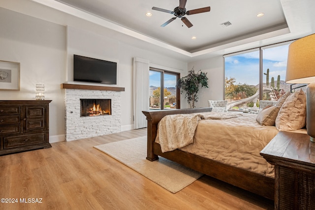 bedroom with a raised ceiling, a stone fireplace, ceiling fan, and light hardwood / wood-style flooring