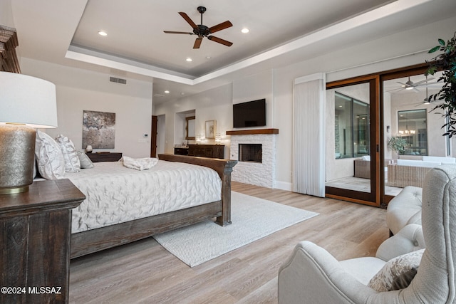 bedroom featuring ceiling fan, a stone fireplace, a tray ceiling, access to outside, and light wood-type flooring