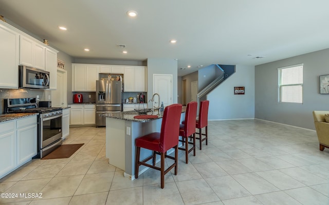 kitchen with white cabinets, stainless steel appliances, a kitchen island with sink, and dark stone counters