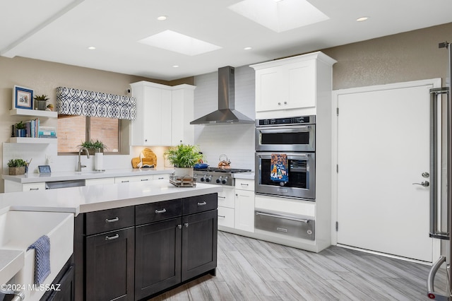 kitchen with wall chimney range hood, a warming drawer, white cabinets, and a skylight