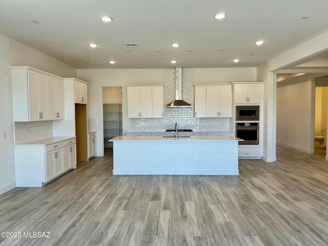 kitchen with wall chimney exhaust hood, a sink, light wood-type flooring, an island with sink, and stainless steel appliances