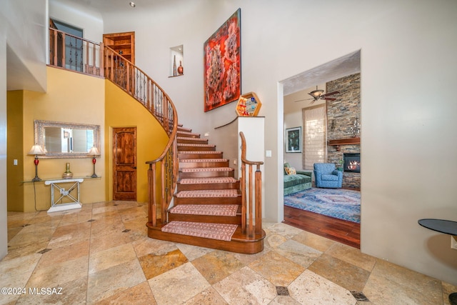 stairs with a stone fireplace, ceiling fan, a towering ceiling, and wood-type flooring