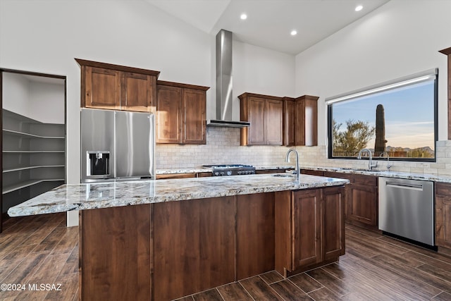 kitchen featuring wood finish floors, an island with sink, a sink, appliances with stainless steel finishes, and wall chimney range hood