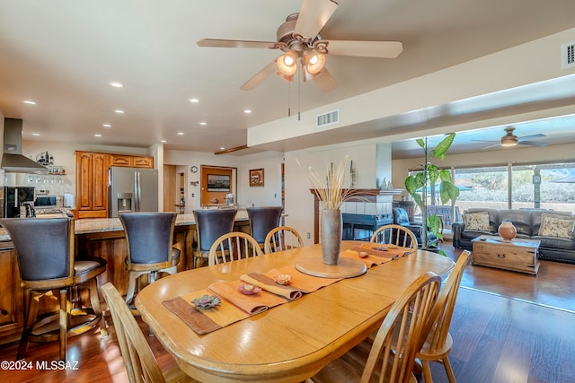 dining area with a tiled fireplace, ceiling fan, and dark wood-type flooring