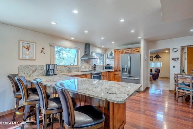 kitchen with wall chimney exhaust hood, kitchen peninsula, wood-type flooring, a breakfast bar, and appliances with stainless steel finishes