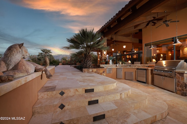 patio terrace at dusk featuring area for grilling, ceiling fan, and exterior kitchen