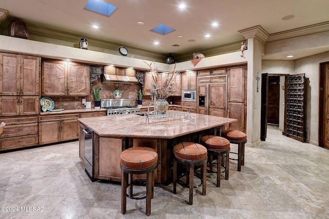 kitchen with built in appliances, wall chimney range hood, a skylight, and an island with sink