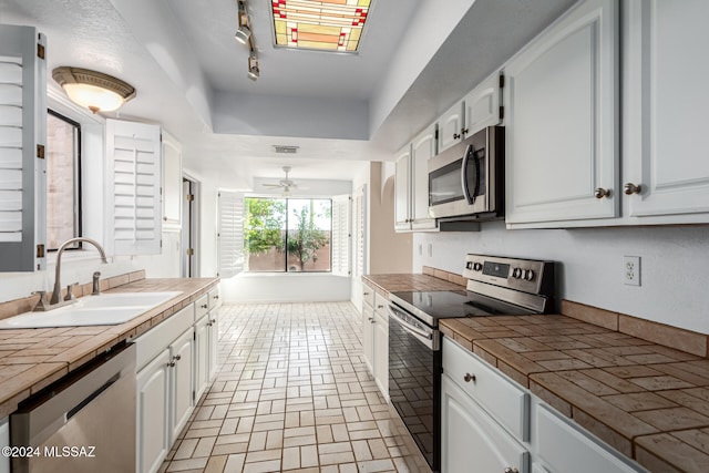 kitchen with stainless steel appliances, ceiling fan, sink, tile countertops, and white cabinets