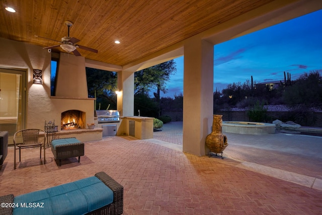 patio terrace at dusk with a large fireplace, grilling area, ceiling fan, and exterior kitchen