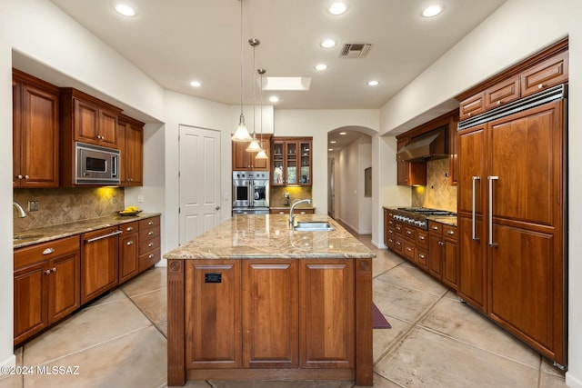 kitchen featuring tasteful backsplash, light stone counters, built in appliances, an island with sink, and decorative light fixtures