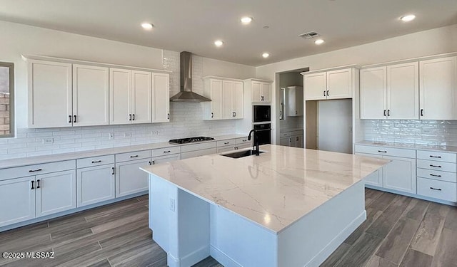 kitchen with wall chimney range hood, gas cooktop, tasteful backsplash, a sink, and an island with sink