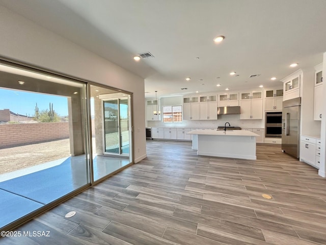 kitchen with tasteful backsplash, white cabinetry, light countertops, under cabinet range hood, and a kitchen island with sink