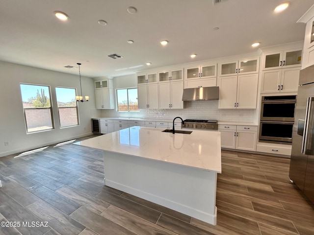 kitchen with white cabinetry, under cabinet range hood, decorative backsplash, double oven, and a sink
