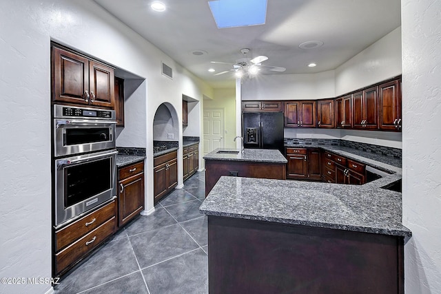 kitchen with dark stone counters, a center island with sink, black fridge, a skylight, and double oven