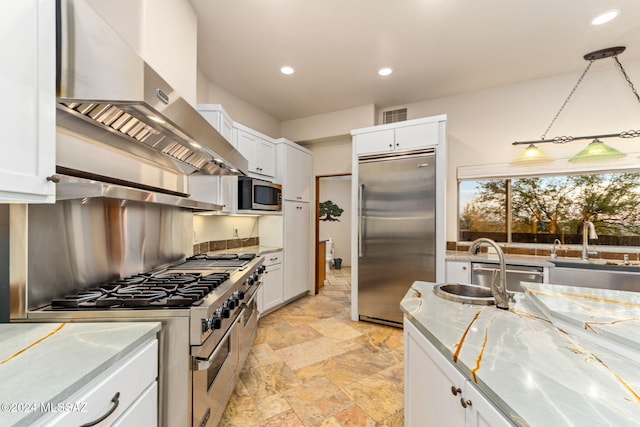 kitchen with light stone counters, wall chimney exhaust hood, premium appliances, white cabinets, and hanging light fixtures