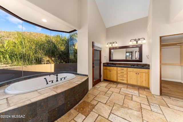 bathroom with high vaulted ceiling, vanity, a relaxing tiled tub, and a wealth of natural light