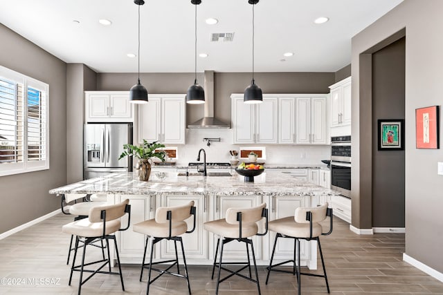 kitchen featuring wall chimney exhaust hood, stainless steel appliances, decorative light fixtures, a center island with sink, and white cabinetry