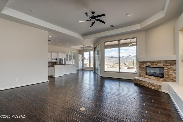 living room featuring dark hardwood / wood-style flooring, a tray ceiling, a stone fireplace, and ceiling fan