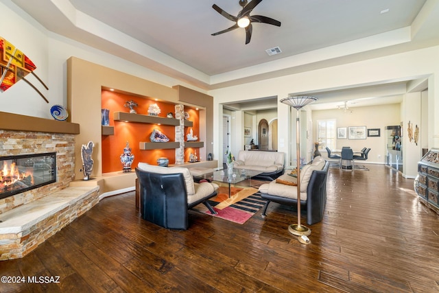 living room featuring a tray ceiling, a stone fireplace, ceiling fan, and dark hardwood / wood-style floors