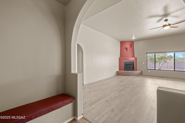 unfurnished living room featuring ceiling fan, a fireplace, arched walkways, baseboards, and wood finished floors