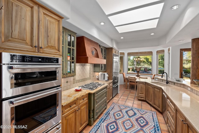 kitchen with sink, stainless steel appliances, light stone counters, custom exhaust hood, and a notable chandelier