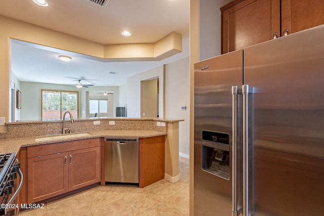 kitchen featuring ceiling fan, light stone counters, appliances with stainless steel finishes, brown cabinets, and a sink