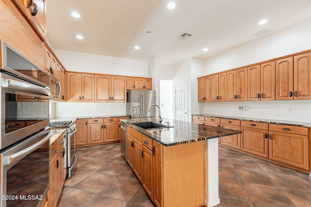 kitchen featuring a kitchen island with sink, dark tile patterned flooring, sink, dark stone countertops, and appliances with stainless steel finishes