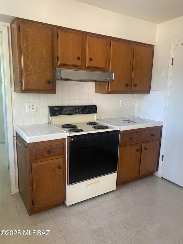 kitchen featuring tile countertops, under cabinet range hood, range with electric stovetop, backsplash, and brown cabinets