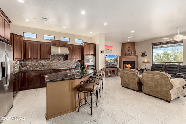 kitchen with stainless steel refrigerator with ice dispenser, sink, a large fireplace, an island with sink, and range hood