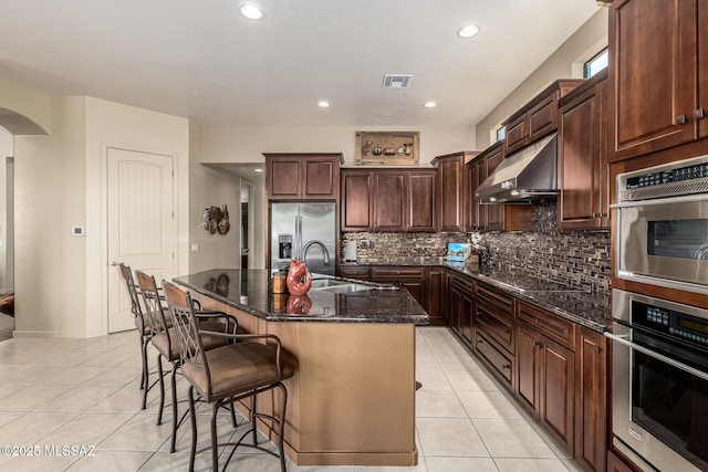 kitchen with light tile patterned flooring, tasteful backsplash, a center island with sink, dark stone countertops, and stainless steel appliances