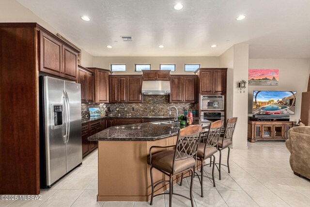 kitchen with dark stone countertops, backsplash, a kitchen bar, stainless steel refrigerator with ice dispenser, and a center island with sink