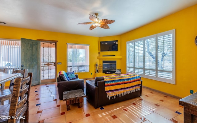 tiled living area featuring ceiling fan, baseboards, and a glass covered fireplace