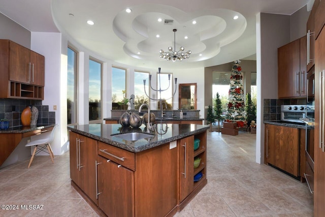 kitchen featuring dark stone countertops, a center island with sink, open shelves, a sink, and a raised ceiling