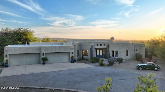 pueblo revival-style home featuring stucco siding, an attached garage, a chimney, and driveway