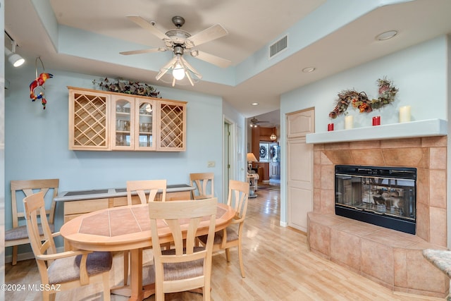 dining room with a fireplace, light wood-type flooring, a tray ceiling, and ceiling fan