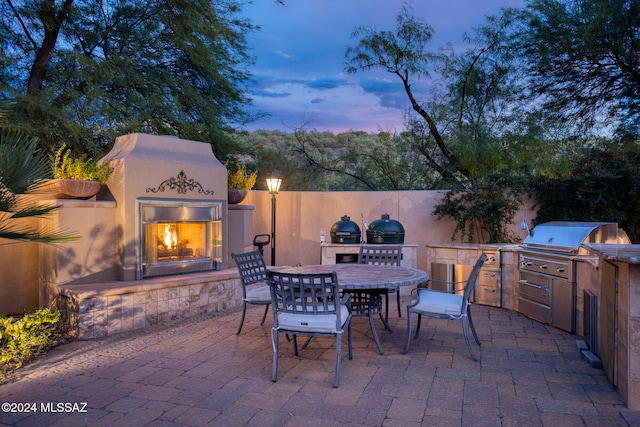 patio terrace at dusk featuring grilling area, an outdoor fireplace, and exterior kitchen