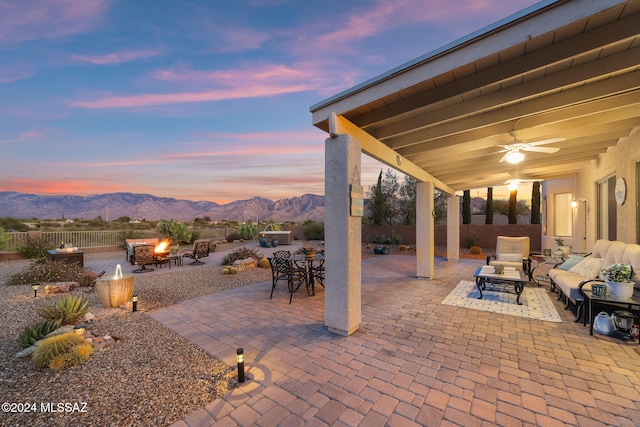 patio terrace at dusk with ceiling fan, an outdoor living space with a fire pit, and a mountain view