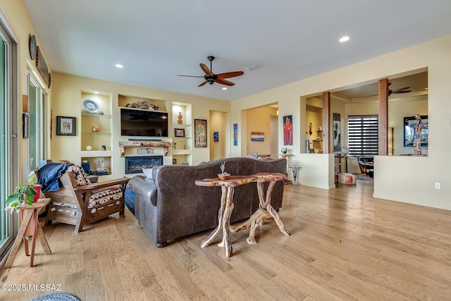 living room featuring light wood-style flooring, visible vents, built in features, a ceiling fan, and a glass covered fireplace