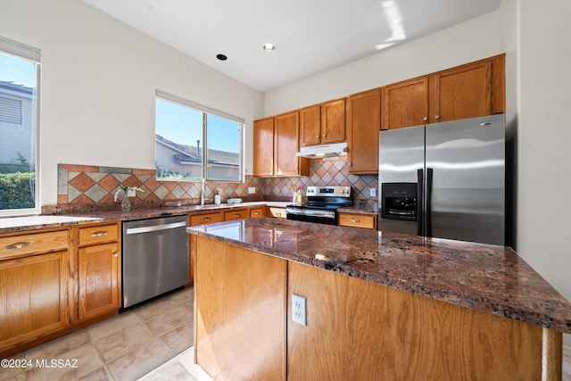 kitchen with brown cabinets, under cabinet range hood, dark stone countertops, backsplash, and appliances with stainless steel finishes